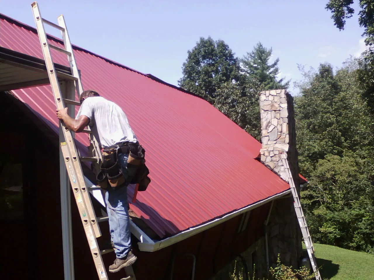 Roofer on a ladder beside a red metal roof and stone chimney — Soffit & Fascia Repair by Roofing Brothers in Brayton